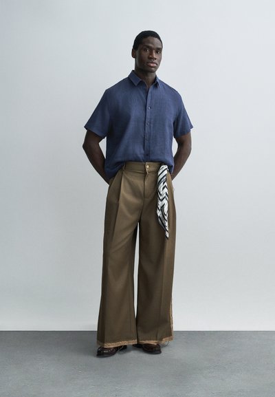 Άντρας που στέκεται μπροστά σε γκρι τοίχο, φοράει μπλε πουκάμισο με κοντά μανίκια και κουμπιά, φαρδιά καφέ παντελόνια και μαύρο-άσπρο μαντήλι με σχέδιο που κρέμεται από την τσέπη.