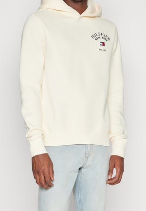 Homme portant un sweat à capuche Hilfiger New York blanc cassé avec logo à l'avant et un jean bleu clair, debout devant un fond blanc uni.