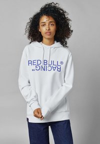 Sudadera blanca con un bolsillo tipo canguro, con una impresión en negrita "RED BULL RACING" en azul. Tejido suave con cuerdas ajustables, corte relajado.