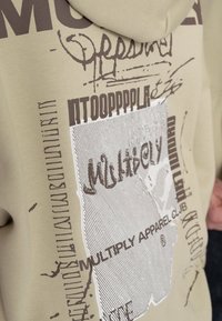 Béžová mikina s hnědým grafickým designem, obsahující abstraktní vzory a text, včetně "Multiply Apparel Club," na měkké, strukturované tkanině.