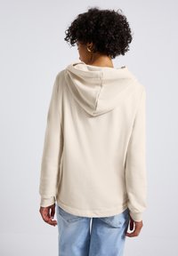 Felpa beige con cappuccio regolabile, polsini a costine e un fit rilassato. Texture morbida visibile da dietro, abbinata a jeans blu chiaro.