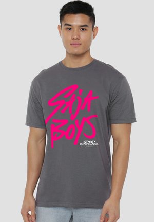 Junger Mann trägt ein graues T-Shirt mit leuchtend pinkem "Saija Boys" Schriftzug und kleinerem "Kpop Demon Hunters" Logo vorne, steht vor einfachem Hintergrund.