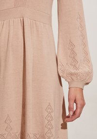 Långärmad rosa stickad klänning med en figurnära överdel och utsvängd kjol. Dekorativa diamantformade utskärningar längs ärmarna och fållen förstärker texturen.