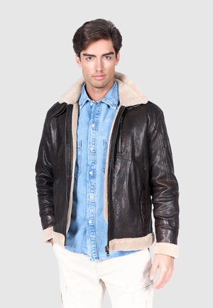 Chaqueta de cuero marrón con cuello y puños de borrego, con cierre de cremallera y bolsillos laterales, llevada sobre una camisa de mezclilla.