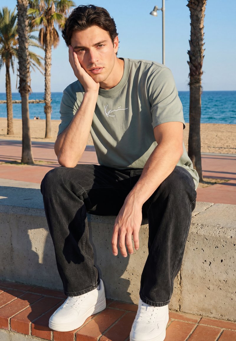 Jonge man in een groen T-shirt en zwarte broek zit op een betonnen richel bij het strand, met zijn hoofd rustend op zijn hand, met de oceaan en palm bomen erachter.