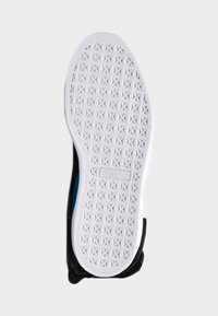 Svart idrottssko med en vit gummisula som har ett strukturerat greppmönster och en blå detalj synlig på sidan.