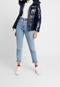 Námořnická lesklá puffer bunda s kapucí, kombinovaná s vysokým pasem světle modrými džíny a bílými teniskami. Červené ponožky jsou viditelné nad botami.