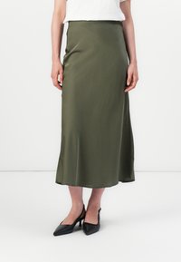En person står klädd i en vit skjorta och en olivgrön satin midi-kjol, kompletterad med eleganta svarta högklackade skor.