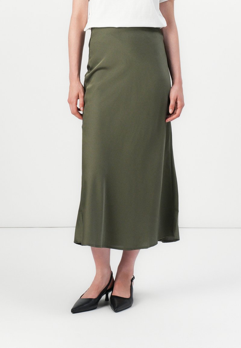En person står klädd i en vit skjorta och en olivgrön satin midi-kjol, kompletterad med eleganta svarta högklackade skor.