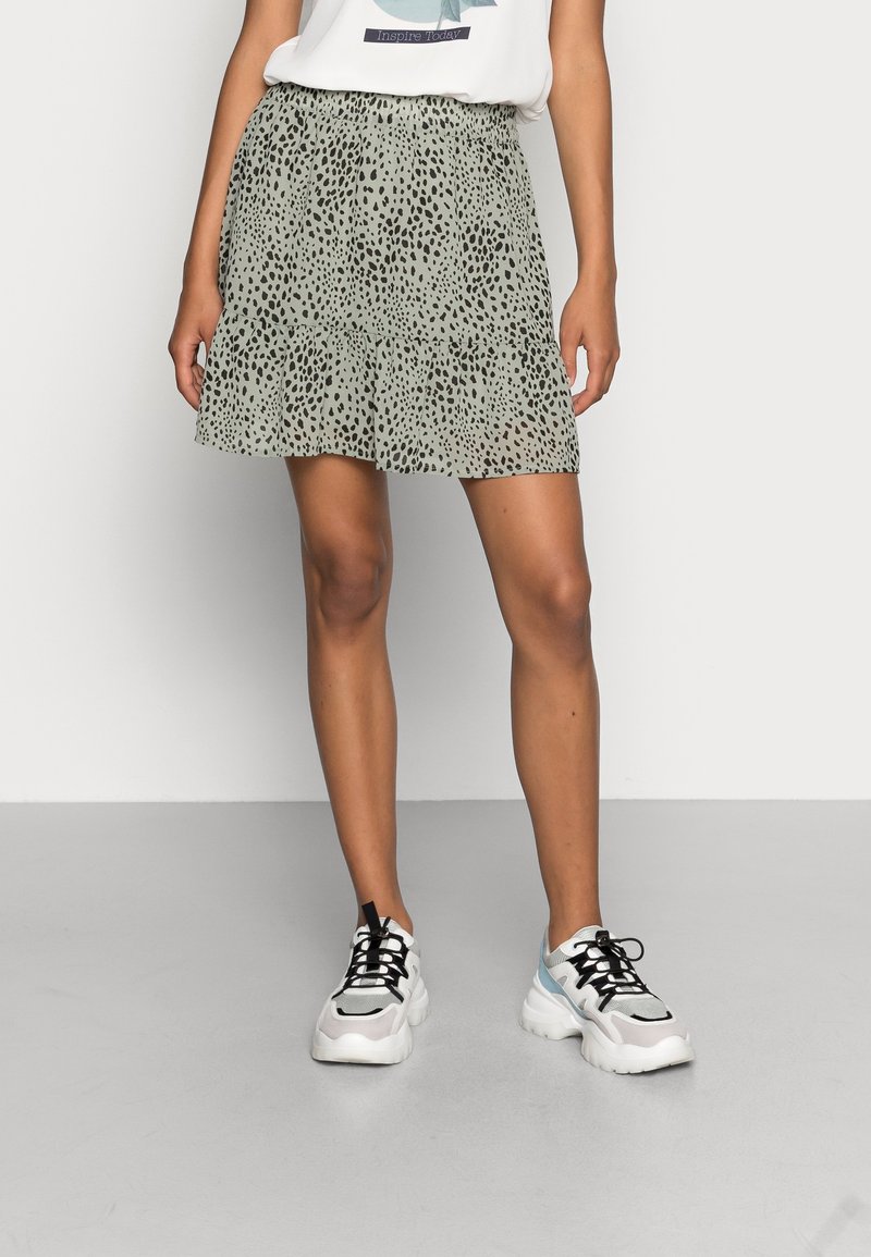 Grön volangkjol med svarta prickar, försedd med elastisk midja. Matchad med kraftiga vita och blå sneakers mot en neutral bakgrund.