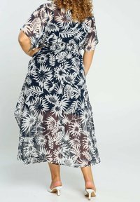 Kvinna som bär en svart och vit blommig genomskinlig midi-klänning, står med händerna på höfterna, lockigt hår och vita klackskor på en enfärgad bakgrund.