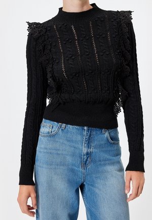 Femme portant un pull en maille noire texturée avec des détails en dentelle et un jean bleu taille haute, debout avec les bras détendus le long du corps.