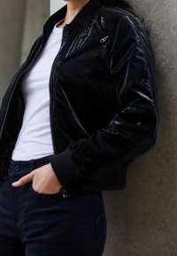 Black bomber jacket with a shiny finish, zip pocket on the chest, ribbed cuffs, and waistband. Worn over a white t-shirt and dark jeans.