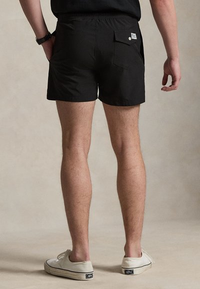 Άνδρας που φοράει μαύρα σορτς με πίσω τσέπη και λευκά αθλητικά παπούτσια, στέκεται με φόντο ένα απλό ουδέτερο φόντο, από πίσω.