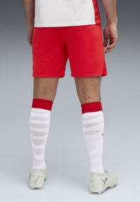 Pantalones cortos deportivos rojos con un diseño ajustado. Medias blancas hasta la rodilla que presentan un patrón texturizado. Botas de fútbol grises con logotipos visibles.