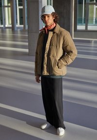 Brun puffjacka med knäppning, i kombination med en grön topp, svarta byxor, vita sneakers och en ljusblå keps, ställd mot en upplyst bakgrund.