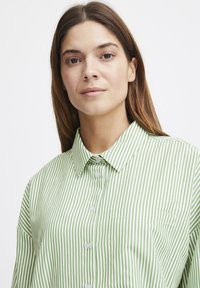 Green and white striped button-up shirt with a collar, featuring long sleeves and a chest pocket. Smooth fabric with a tailored fit.
