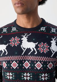 Homem com um suéter de Natal azul-marinho com renas brancas e padrões de flocos de neve vermelhos e brancos.