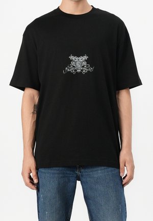 Mand iført en sort T-shirt med en lille detaljeret hvid grafik og tekst i midten, kombineret med blå jeans.