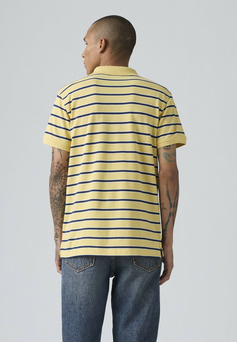 Homme tatoué portant un polo à manches courtes rayé jaune et bleu marine et un jean bleu, vu de dos devant un fond uni.