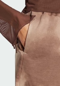 Brown long-sleeve top with sheer, textured sleeves and a waist pocket on beige, loose-fitting pants made of a soft, smooth fabric.