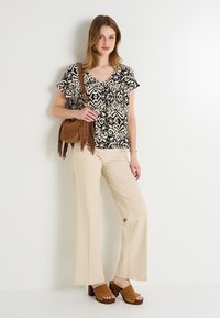 Top de manga corta con un estampado negro y crema, pantalones beiges acampanados, zuecos de plataforma marrones y un bolso cruzado de ante marrón con flecos.