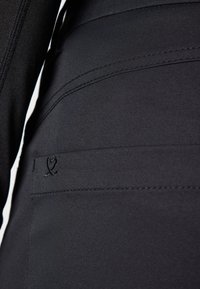 Juodas audinys arti, matomas siuvimas ir mažas, išsiuvinėtas logotipas. Tekstūra lygi, su matinio paviršiaus apdaila.