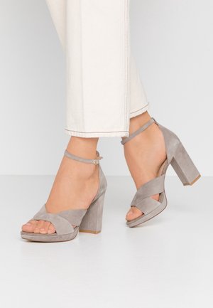 Grå ruskinds sandaler med blokhæle, krydsede stropper over tæerne og en tynd ankelrem. Har en kraftig hæl og struktureret overflade.