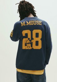 Tummansininen huppari, jossa on keltaisia graafisia elementtejä; takana tekstit "M. MOUSE" ja numero "28" sekä kuva Mickey Hiirestä.