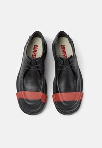 Zapatos de cuero negros que presentan una amplia banda de goma roja en la zona de los dedos, con suelas texturizadas y una plantilla acolchada beige etiquetada como "CAMPER."