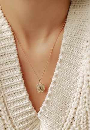 Goldene Halskette mit einem runden Anhänger, der ein erhabenes Design und winzige Verzierungen aufweist, gelegen auf einem strukturierten, cremfarbenen Pullover.