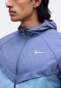 Gros plan d'une personne portant une veste zippée bleue Nike avec capuche, montrant le côté gauche du visage et le haut du torse.