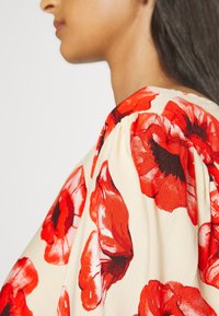 Blusa floreale con motivi di fiori rossi su sfondo beige, dettagli con maniche a sbuffo e tessuto dalla consistenza liscia.