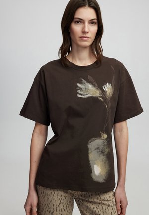 T-shirt di cotone marrone con un design floreale in colori chiari sul davanti, abbinato a pantaloni beige a fantasia. Vestibilità casual, maniche corte.