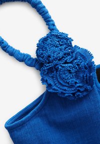 Top de tela azul con tirantes texturizados y fruncidos; acento floral de tela esponjosa en capas; acabado suave en el cuerpo.