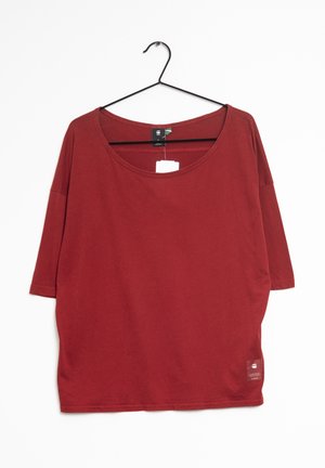 Luźna czerwona koszula z krótkim rękawem i okrągłym dekoltem zawieszona na czarnym metalowym wieszaku na białym tle.