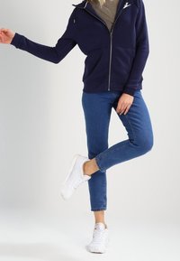 Námořnická mikina na zip s minimalistickým logem, v kombinaci s světle modrými džíny a bílými teniskami. Disponuje volným střihom a hladkým materiálem.