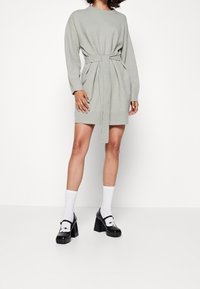 Abito grigio lavorato a maglia con taglio rilassato, maniche lunghe, cintura in vita e orlo testurizzato. Abbinato a scarpe con plateau nere e calzini bianchi.