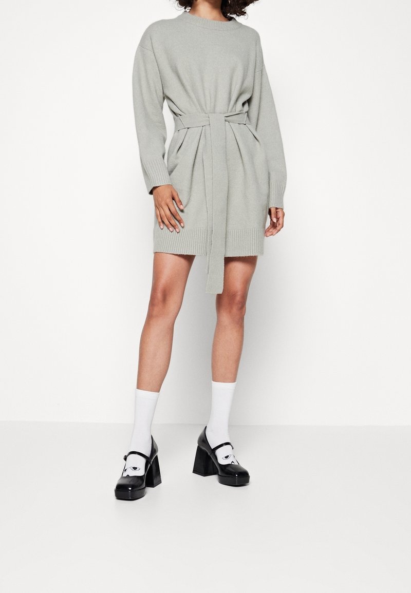 Abito grigio lavorato a maglia con taglio rilassato, maniche lunghe, cintura in vita e orlo testurizzato. Abbinato a scarpe con plateau nere e calzini bianchi.