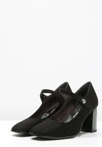 Czarne zamszowe szpilki Mary Jane z zaokrąglonym noskiem, średniej wysokości słupkiem i regulowanym paskiem z metalowym akcentem.