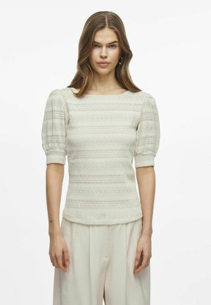Haut en tricot ivoire avec des manches courtes bouffantes, présentant un motif rayé texturé et un décolleté bateau, porté avec un pantalon léger à coupe décontractée.