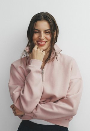 Lächelnde Frau mit dunklem Haar trägt ein rosa Sweatshirt mit Reißverschluss und stützt ihr Kinn mit der Hand vor einem schlichten hellen Hintergrund ab.