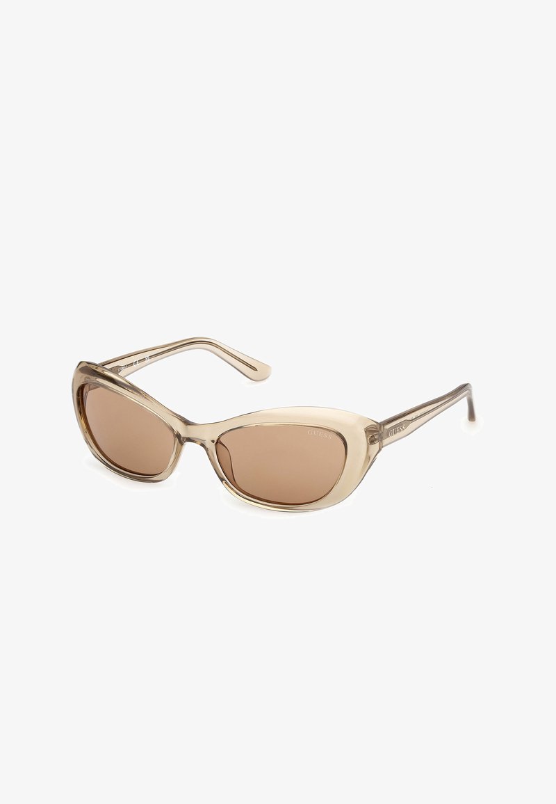 Lunettes de soleil beige translucides à monture œil de chat avec des verres teintés marron et des branches fines, posées à plat sur une surface blanche.
