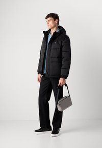 Jovem vestido com uma jaqueta acolchoada preta, uma camisa de denim clara, calças pretas e ténis pretos, segurando uma mala cinza de ombro contra um fundo branco.