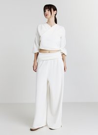 Jeune femme aux cheveux bruns en queue de cheval basse, portant un crop top à manches longues blanc et un pantalon large taille haute, sur fond uni.