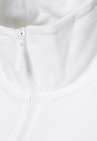 Zbliżenie na biały materiał ubrania z pionowym suwakiem częściowo zasuniętym przy kołnierzu.