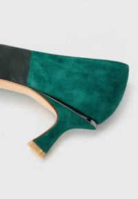 Decolleté verde in suede con punta affusolata e suola nera e beige, caratterizzato da un tacco sottile di altezza media e una superficie liscia.