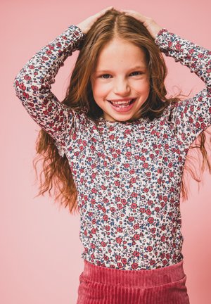 Langsleeve, aansluitend top met een witte basis met rode en blauwe bloemmotieven, gecombineerd met een roze corduroy rok. Achtergrond is roze.