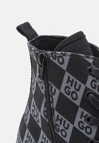 Must-valget musta ja halli ruuduline pahklääts, mis on valmistatud tekstiilist, millel on tõmblukk ja paelad. Saabas sisaldab brändi logo detaile ja tekstuurset pinda.