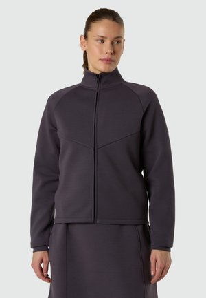 Femme portant une veste zippée gris foncé à col haut et une jupe assortie, debout devant un fond clair uni, regardant vers la droite.